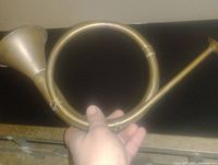 Hand holding the brass French style bugle against a darker background, showing the coiled circular design, the flared bell, and the mouthpiece end.
