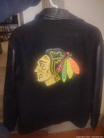Back view of the black jacket showing the large colorful embroidered Chicago Blackhawks logo on the wool body with black leather sleeves.
