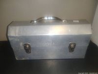 Front view of aluminum lunch box with two metal latches and handle on top. Shows stamped identification marks.