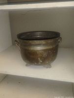 Front view of a solid brass round flower planter pot with two small handles on the sides and three ornate feet, placed on a shelf.