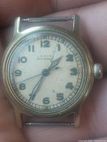 Front face of the wristwatch showing dial with hour and minute hands, winding crown, and slightly worn white dial with Arabic numerals.