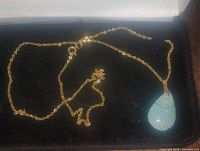 Gold plated chain necklace with a light blue speckled teardrop pendant resting on a black background.