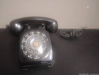Front angled view of the vintage black rotary dial telephone on a black surface showing handset, dial and coiled cord.