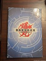 Front cover of Bakugan Battle Brawlers card binder showing series logo and blue tech design background with wear and creases