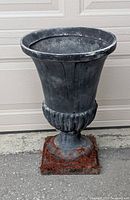 Front angle view of a tall cast iron garden urn with flared top, fluted detailing near base, and square metal base with rust.