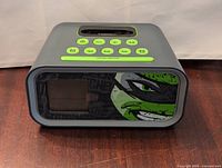 Front view of the gray TMNT alarm clock showing turtle graphic, digital screen and green control buttons