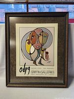 Framed graphic art print showing the full artwork, visible details of the frame and matting. The print features abstract dual faces in muted earth tones.