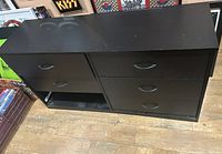 Black bedroom dresser showing five out of six drawers present, missing one drawer on bottom left, photographed on a wooden floor with some background items