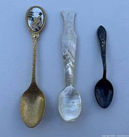 Photo of three collectible spoons: one decorative brass-toned with silhouette, one mother-of-pearl with floral engravings, and one small dark metal spoon with markings.