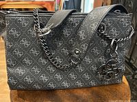 Side view of a black Guess purse with a grey repeated logo pattern, silver-tone chain handles intertwined with black leather handles, and a silver Guess logo charm.