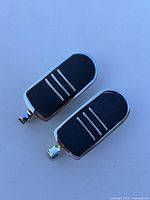 Pair of motorcycle highway foot pegs on a white background showing chrome edges and black rubber tops with three horizontal silver lines.