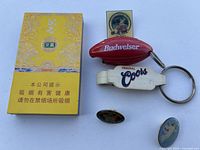 Photo of yellow Japanese cigarette box, Budweiser blimp keychain, white Coors bottle opener keychain, and three assorted pins