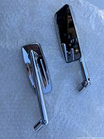 Two chrome motorcycle mirrors laid out on white textured fabric, showing front reflective surfaces and adjustable arms with mounting ends.