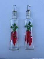 Photo showing two clear glass soap dispensers, each about 10 inches tall with a red chili pepper and green leaf design, topped with metal pump dispensers.