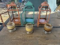 Three outdoor metal lanterns and three small glass candle holders with wax on a wooden surface, outdoors.