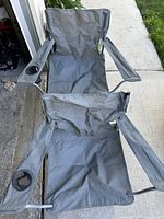 Two gray fabric foldout chairs with metal frames, both with armrests and cup holders on the right armrest, positioned outdoors near a sidewalk and garage.