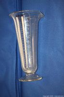 Full side profile of clear glass apothecary measuring beaker, showing graduated etched lines with numerals from 1 to 8 ounces and flared spouted rim. The foot shows some wear and small chip visible.