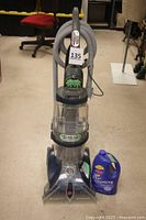 Full upright carpet cleaner with hose and new gallon bottle of cleaning fluid placed on floor beside it