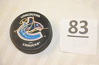Top-down photo of an NHL hockey puck featuring the Vancouver Canucks logo and a gold signature on the top surface alongside an auction lot number 83 card.