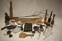 Full assortment of wooden and metal kitchenware including wooden stirrers, metal scoops, wire beaters, molds, and rug beater arranged on white background.