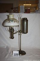 Full view of late 1800s single arm brass kerosene oil student lamp with adjustable height, showing base, reservoir, arm, burner, and floral painted shade with chimney.