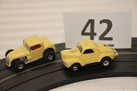 Side view of two yellow vintage Aurora slot cars positioned on a segment of black track, showing the Hot Rod Coupe with red seat and Willys Gasser with number 3 decal.
