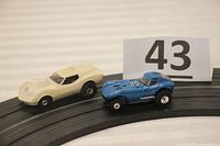 Two Aurora HO scale slot cars on black slot car track, seen side by side, blue T-Jet Cheeta and off-white T-Jet Corvette Mako Shark, with lot number card.