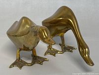 Front and side view of two vintage brass duck figurines with shiny golden finish, showing their detailed feet and heads, on neutral background.