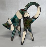 Front side view showing colorful raku pottery cat with looping tail and patchwork glaze of teal, white, brown, and olive green.