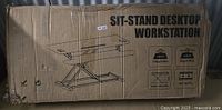 Box packaging with printed diagram and specifications of the sit-stand desktop workstation showing the height adjustable X-frame design and dual-tier desktop surfaces.