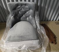 Front view of grey velvet Bellcrest accent chair top with button tufted backrest and rounded cushion, partially wrapped in plastic, with detached wooden legs and hardware to the side.