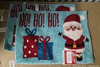 Top view of two Holiday Time bath rugs with festive holiday designs of Santa and gifts on a light blue background