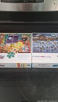 Photo showing two puzzle boxes side-by-side: one with an indoor workshop illustration, the other a winter village skating scene. Both labeled as Buffalo 1000 piece puzzles.