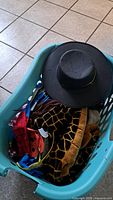 Basket containing multiple children's costumes and accessories, showing a black wide-brim hat on top and various colorful costume fabrics beneath.