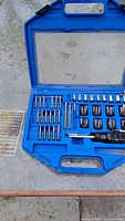 Open blue plastic case containing assorted screwdriver and socket bits arranged in slots. Visible dirt and wear marks on case.
