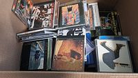 Box filled with multiple jewel case music CDs stacked loosely showing album covers.