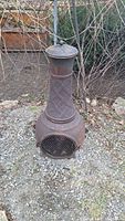Full front view of the tall cast iron chiminea placed outdoors on gravel, showing mesh door, lid with knob, and diamond pattern on chimney body.
