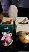 Four collectible Christmas balls and a white salt statue on a black surface. Three balls are outside boxes; one ball inside a Regal box. Coca-Cola ribbon on one ball and cherubic gold floral designs visible.