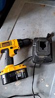 Yellow and black DeWalt drill with attached 18V battery lying on a dirty gray surface next to a black charging unit with power cord.
