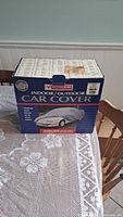Box of Motomaster indoor/outdoor car cover on table with lace tablecloth, showing front of box with product name and image
