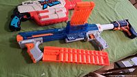 Photo of Nerf Mega XL Big Rig blaster and Nerf Rampage blaster on green surface with orange ammo clips and darts.