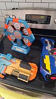 Nerf electronic target alongside a tan and orange blaster and a large blue blaster on a black stove