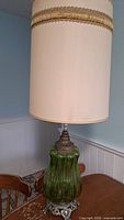 Full view of one lamp showing green textured glass body, ornate metal base, and tall beige fabric shade with gold trim.