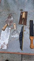 Photo showing five patching tools laid out on a concrete surface including trowels, scrapers, a brush, and sanding block.