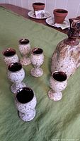 Photo of the six small pottery cups and the pitcher on a green tablecloth, showing the speckled glaze and dark brown rims