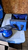 Top angle showing the full Simoniz buffer, polishing pad, and nearby blue carrying case on plastic container lid.