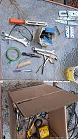 Photo of assorted hand tools and door hardware laid out on glass surface including wrenches, locking pliers, drill bits, tape measure, and a box of screws and nails.