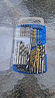 Blue plastic drill bit case with clear cover open showing various sizes of drill bits in gold, silver, and black finishes.