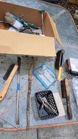 Top perspective showing cardboard box with small metal tools and several wire brushes and metal cleaning tools laid out on a glass table outdoors.