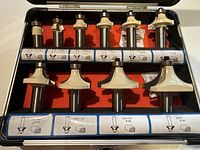 Top view of the 12 carbide router bits in black case with red lining. Each bit has a white cutting head and a 1/2 inch shank. Illustrations and measurements of each bit profile are shown on the case interior label.